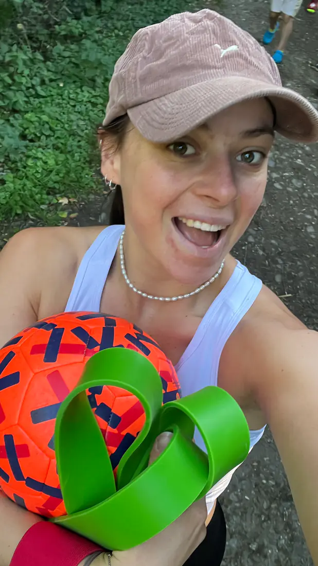 Eine Frau hält lachend einen Ball und grüne Gymnastikbänder in die Kamera, dahinter ein Waldweg.