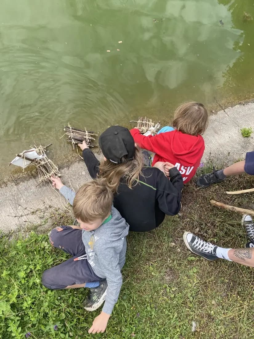 Kinder liegen am Ufer und schieben ein selbst gebautes Floß aus Ästen ins Wasser.
