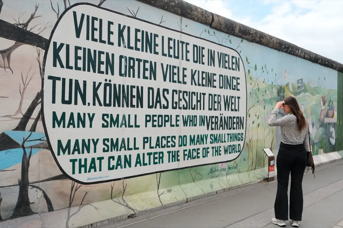 Judith steht vor einer bemalten Mauer mit dem Spruch, dass viele kleine Leute an vielen kleinen Orten das Gesicht der Welt verändern können.
